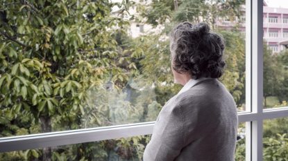 senior woman with gray hair gazing out a window at the trees, grieving