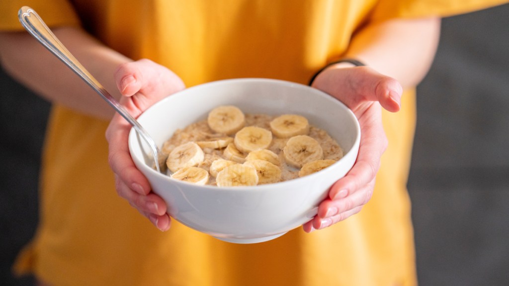 A close up of a woman in a yellow top holding a white bowl of cereal topped with a banana, which is a potassium-rich food