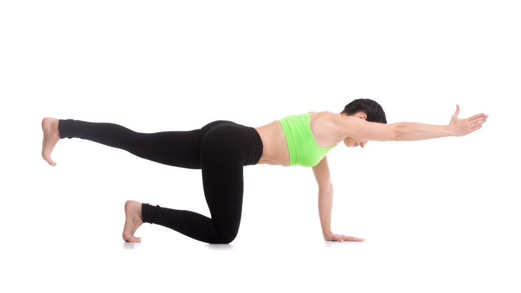 A woman with short dark hair wearing black pants and a green sports bra doing bird-dog yoga exercises for uneven hips