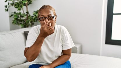 mature woman sitting on white bed holding her nose, bad breath