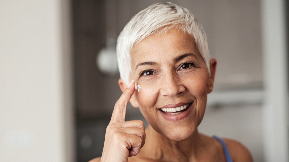 Woman with gray hair and smiling with her finger pointed at the corner of one of her eyes. She's applying a cream after learning about the best crow's feet remedies