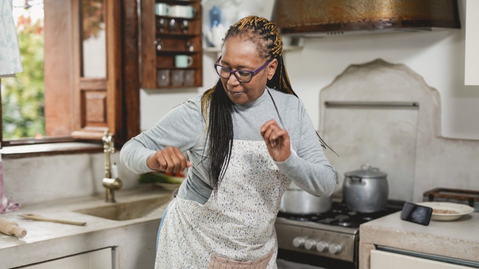 mature woman with glasses and braids dancing in her kitchen, a way to reduce osteoporosis risk