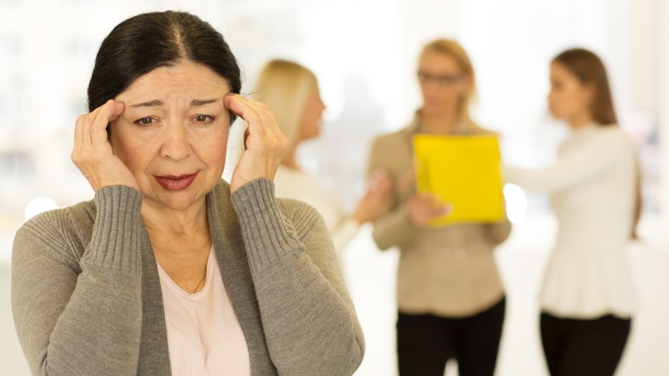 Senior woman having headache at work rubbing her temples her colleagues talking on the background copyspace stressing out stress depressing painful aching migraine overworking retirement.
