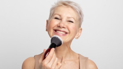 Beauty portrait mature woman applying blush with a makeup brush over grey background.