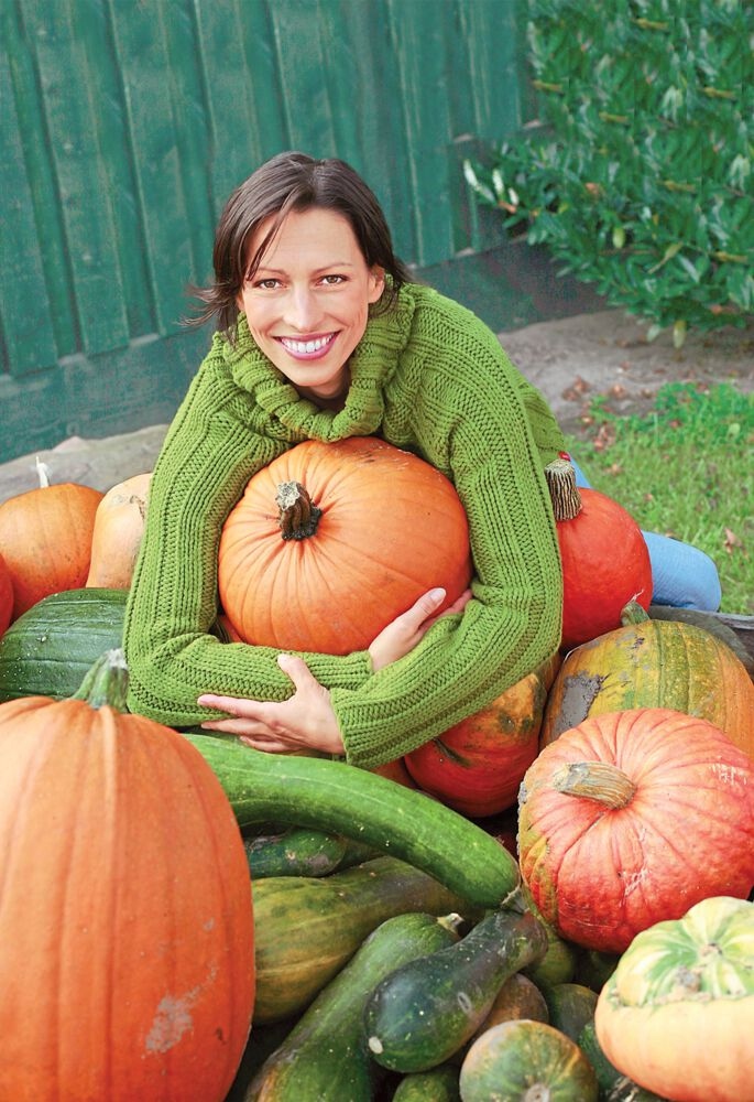 woman hugging a pumpkin with a green stem, next to gourds and pumpkins with pale stems
