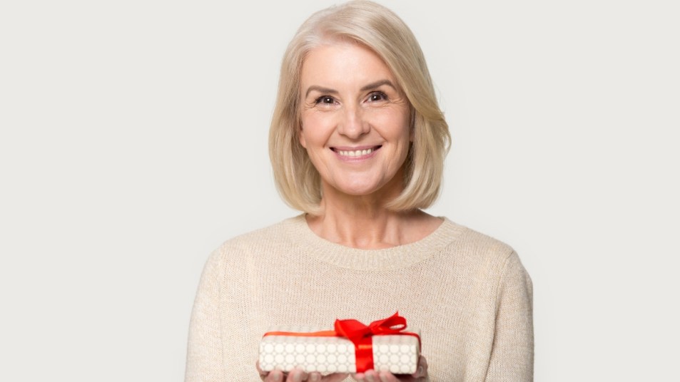 Attractive aged blond woman looks at camera holding presents gift box red bow package feels happy studio head shot isolated on grey background. Life events celebration congratulation love care concept