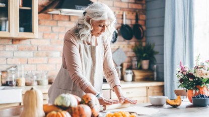 Aged lady with long loose grey hair kneads dough among fresh products at home.