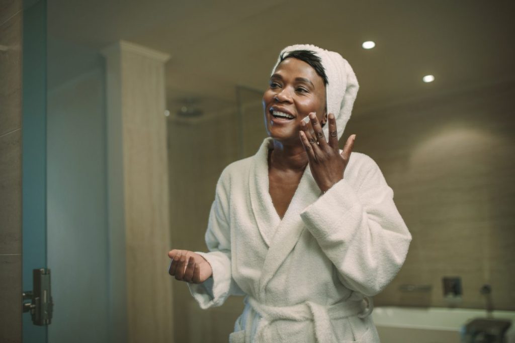 Smiling african woman applying cosmetic cream on face standing in the bathroom. Reflection in mirror of female putting body lotion cream on her face.