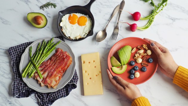 Woman's hands holding plate of avocado, nuts, and berries, and other keto food like bacon, cheese, eggs, and vegetables on table