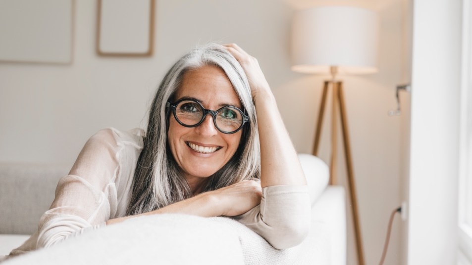 A woman with long grey hair and black glasses sitting on a white couch to strengthen her memory