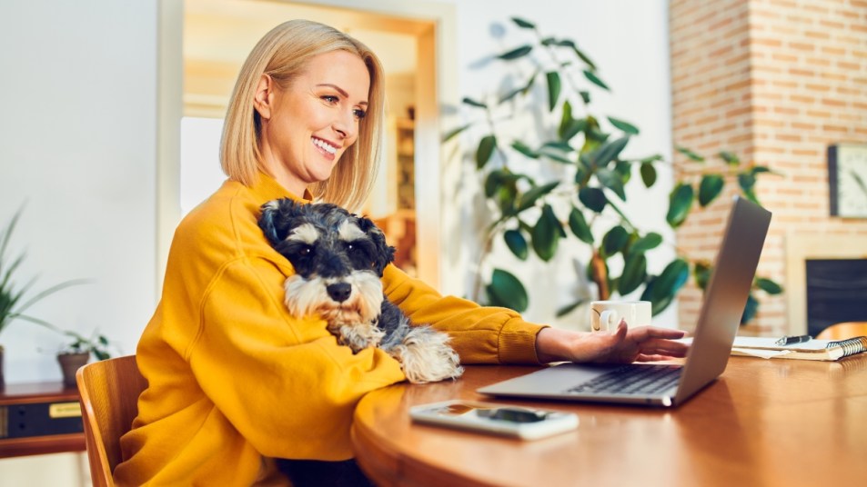 Smiling middle aged woman holding dog using laptop while at home office