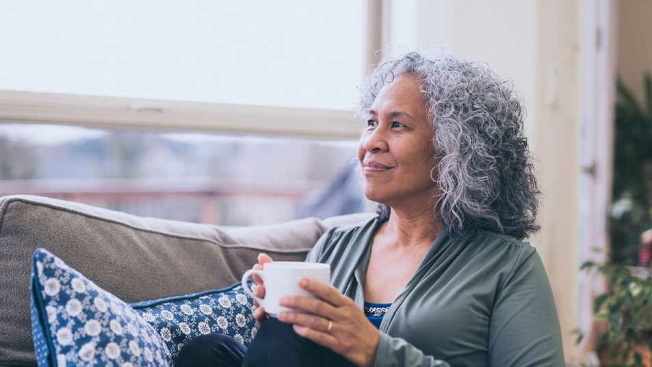 Mature Hawaiian woman with gray curly hair gets a mood-boosting moment while sitting on her couch with a cup of tea in her hands, smiling faintly and looking out the window.