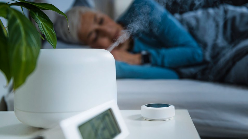 What does a humidifier do? Woman sleeping in bed in background, with humidifier on nightstand