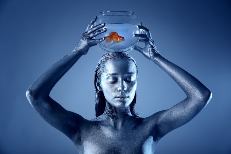 Pisces woman painted in blue holding a bowl with a fish swimming inside of it.