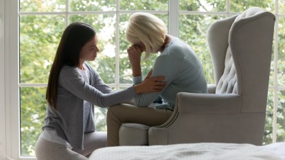 Near panoramic window crying elderly mom sit on armchair, near sit her grown up daughter comforting her in difficult life period give support, share pain at divorce, showing attention and care concept