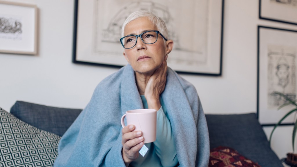 A woman with short grey hair and glasses holding her sore throat while wrapped in a blanket holding a mug of tea