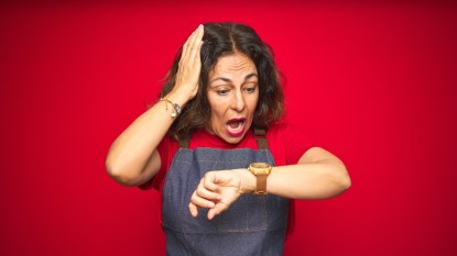 Middle age senior woman wearing apron uniform over red isolated background Looking at the watch time worried, afraid of getting late