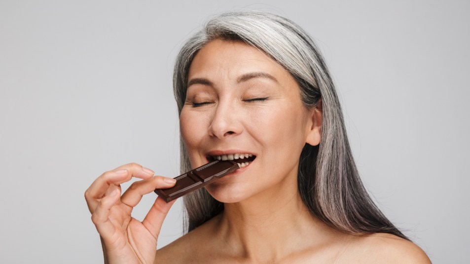 Portrait of mature woman with long gray hair standing over gray background eating chocolate with eyes closed