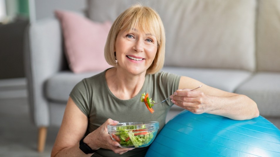 Senior woman with fitness ball eating fresh vegetable salad at home