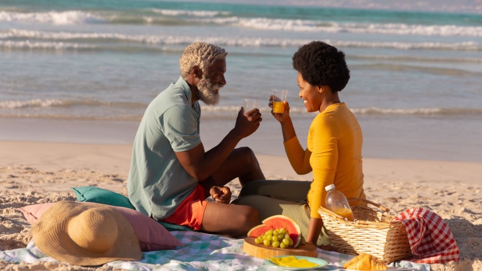 African american couple toasting juice while sitting with food on blanket against sea at sunset. happy, nature, unaltered, love, togetherness, retirement, picnic, enjoyment and holiday concept.