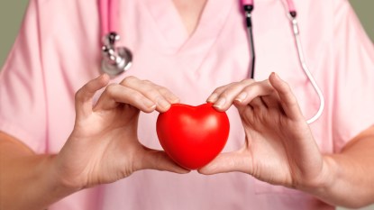A close up of a woman doctor in pink scrubs with a stethoscope holding a red heart object