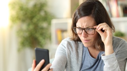 Adult woman with eyesight problems wearing eyeglasses reading on smart phone sitting on a couch at home
