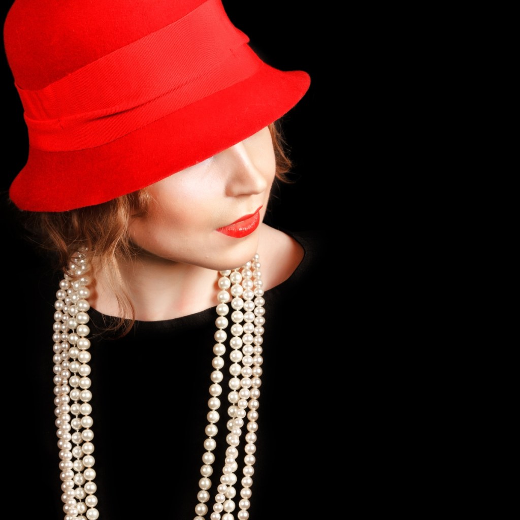 Woman in red cloche hat with red lipstick, pearls, and black dress on black background