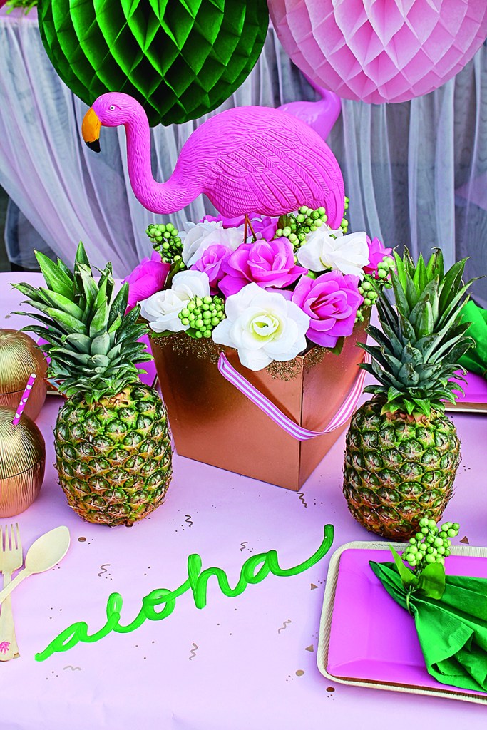 Centerpiece made up of white and pink flowers, a plastic pink flamingo and a couple of pineapples on a table