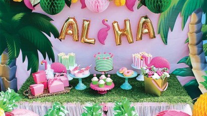 Luau party table with decorations and palm trees, desserts and a balloon sign that says ALOHA is hung behind the table