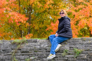 An adult woman sits atop a stone wall in denim jeans, sneakers, and a puffer jacket amid a fall outdoor landscape.