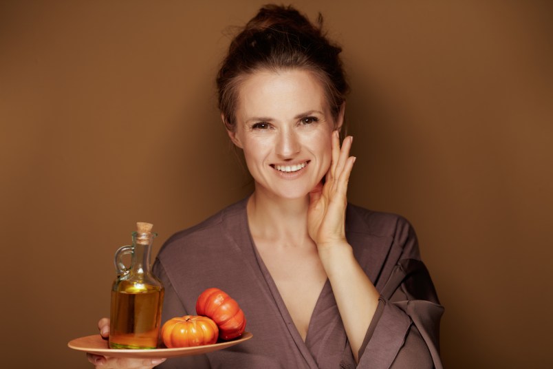 Portrait of happy elegant middle age woman in a bathrobe showing pumpkin oil and touching face on bronze background.