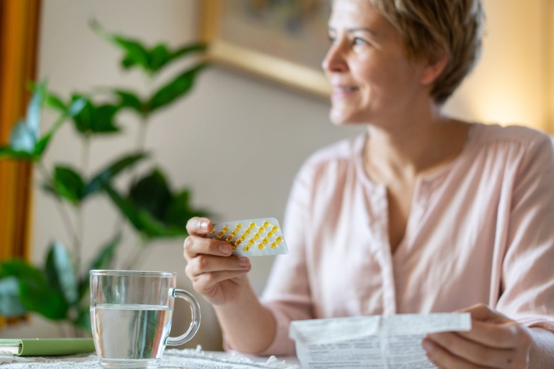 A smiling woman is holding a blister packet of pills and the leaflet, with a drink of water ready on the table sitting at home