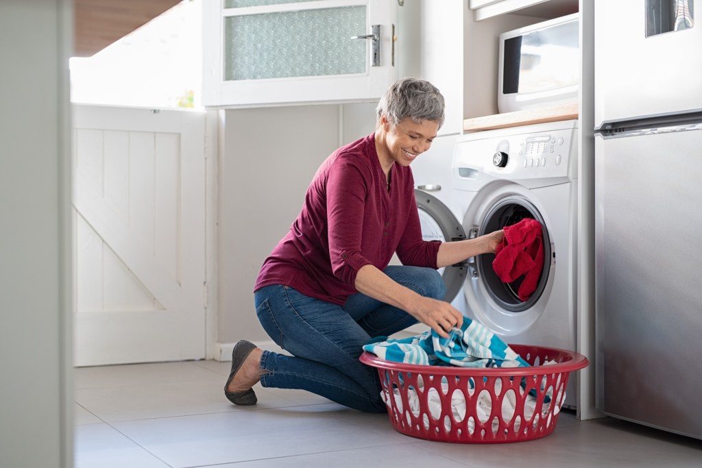 Happy senior woman loading dirty clothes in washing machine.  