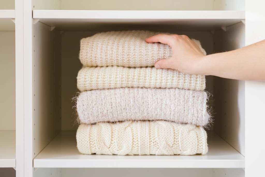 Young adult woman hand taking folded knitted light sweater from shelf in white opened wardrobe at room. (how to fold a sweater)