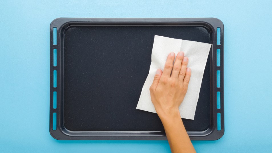 woman hand wiping dark black oven tray with dry white paper napkin on light blue table background. (How to Deep Clean Baking Sheets)