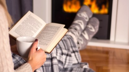 Best loneliness books: featured image that shows a women snuggled up by a fire with a cup of coffee and a book