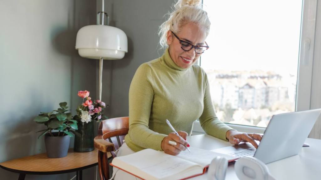 Happy mature woman writing notes and using laptop