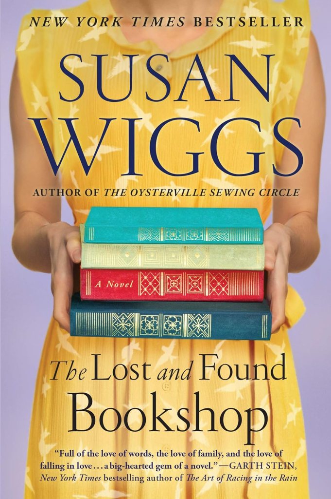 Found Family Trope: The Lost and Found Bookshop by Susan Wiggs book cover shows the torso of a woman in a yellow dress holding a stack of colorful hardcover books