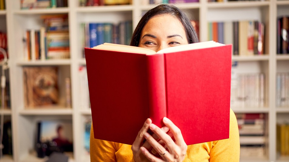 Funny Books feature image of a woman laughing and smiling behind a red hardcover book that she is holding, a bookcase is behind her