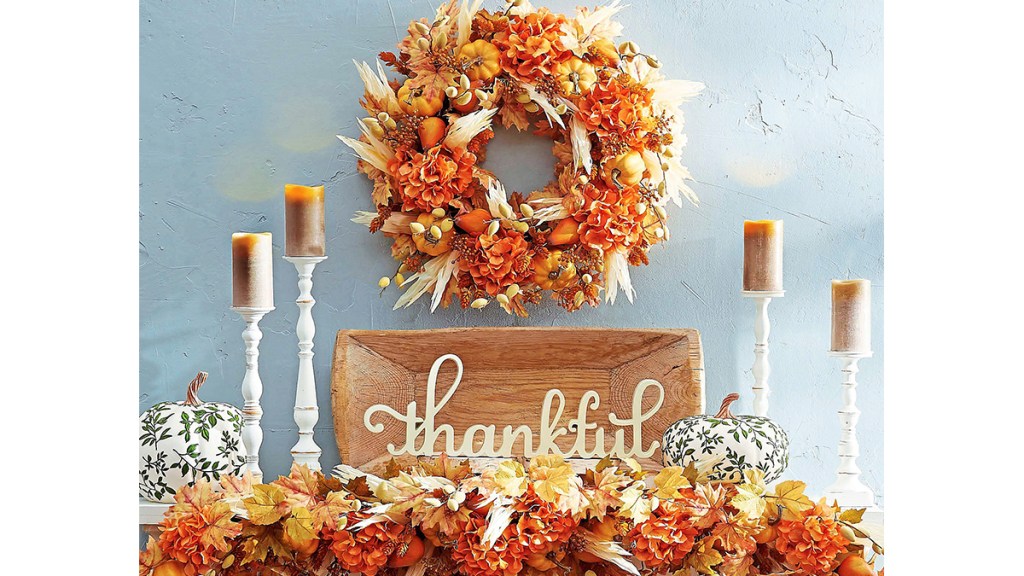 Fall mantel decor ideas: Fireplace mantel or floating shelf decorated cottage style with an orange leafy wreath and garland, white candlesticks, wooden tray and wooden cutout "thankful" sign.