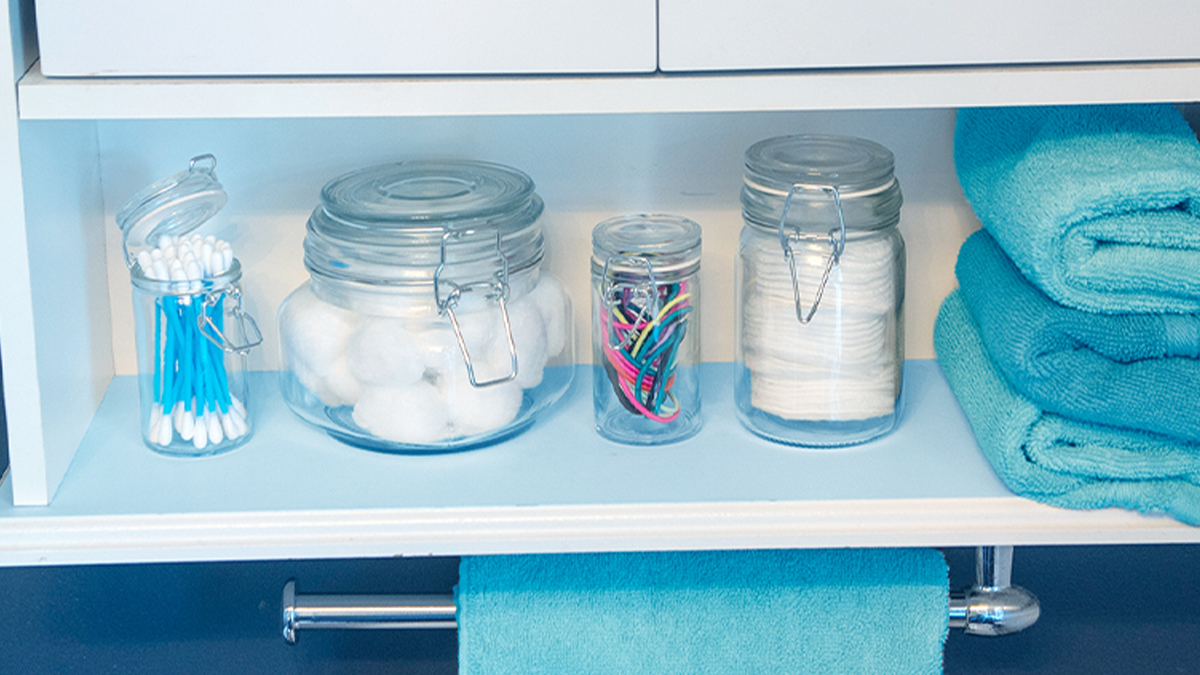 toiletries like cotton swabs, cotton balls and hair ties in empty candle jars on a bathroom shelf with towels
