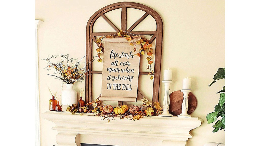 Fall mantel decor ideas: Cream fireplace mantel decorated in a rustic yet refined style with a brown wooden window frame, autumn-themed paper sign, vases filled with leafy branches and pumpkins
