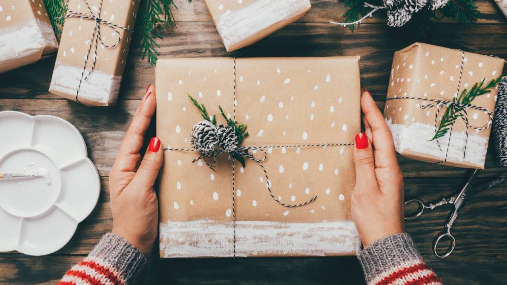 How to wrap a book: Woman´s hands wrapping Christmas presents on brown paper decorated with painted snow, fir branches and pinecones on a rustic wooden board.