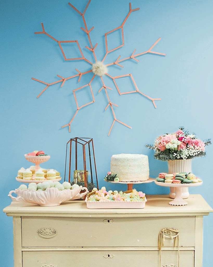 Bar cart decor idea featuring a winter-themed wooden tea cart topped with platters of sweet treats and positioned in front of a large snowflake wall accent made from wooden craft sticks