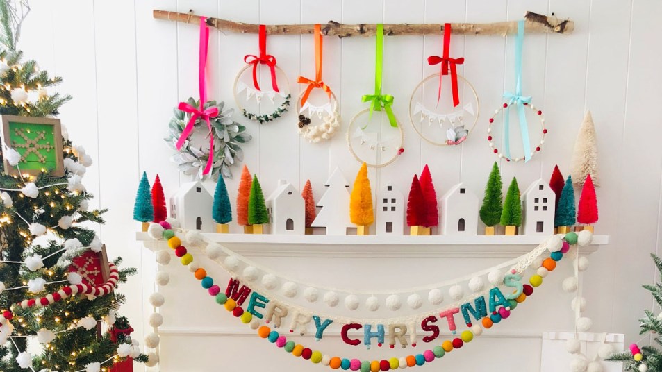 Christmas Mantel Ideas: Colorful boho-style mantel featuring pompom garlands, ribbon-tied embroidery hoops hung on a branch, sisal trees and white mini decorative houses on a white mantel