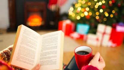 Holiday books: Featured image that shows a woman snuggled up with a book and coffee by a Christmas tree
