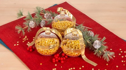 DIY noisemakers made by filling clear plastic ornaments with gold bead necklaces arranged on a red placemat on a wooden tabletop surrounded by evergreen and berry sprigs and extra beads