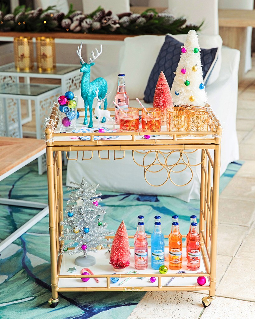 Bar cart decor idea featuring a whimsical gold bar cart with colorful bottled drinks, mini Christmas trees, reindeer, baubles and glassware arranged across both tiers of the cart