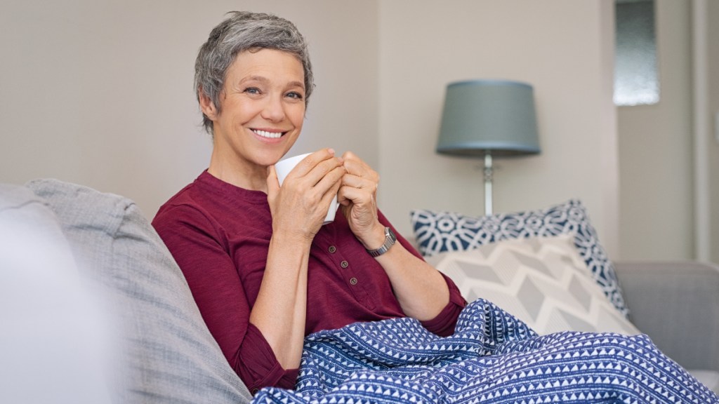 A grey-haired woman in a burgundy sweater drinking chaga tea while under a blanket on the couch