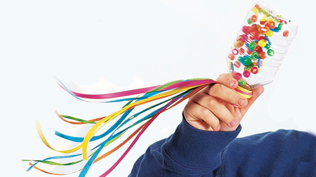 Child's hand holding a DIY noisemaker made by filling a small plastic water bottle with colorful pony beads and buttons and tying ribbon onto the cap